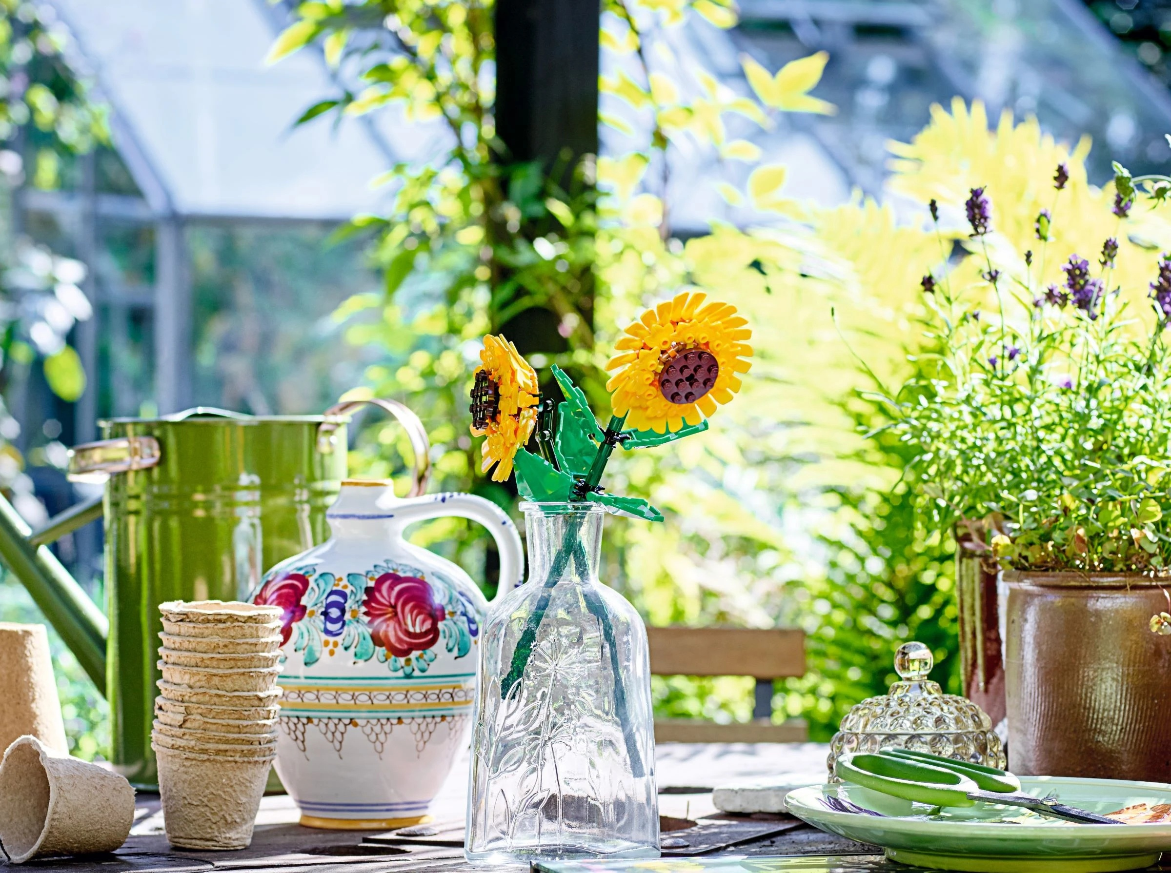 Stół ogrodowy z dwoma żółtymi słonecznikami LEGO w szklanym wazonie, zieloną konewką i doniczkami.