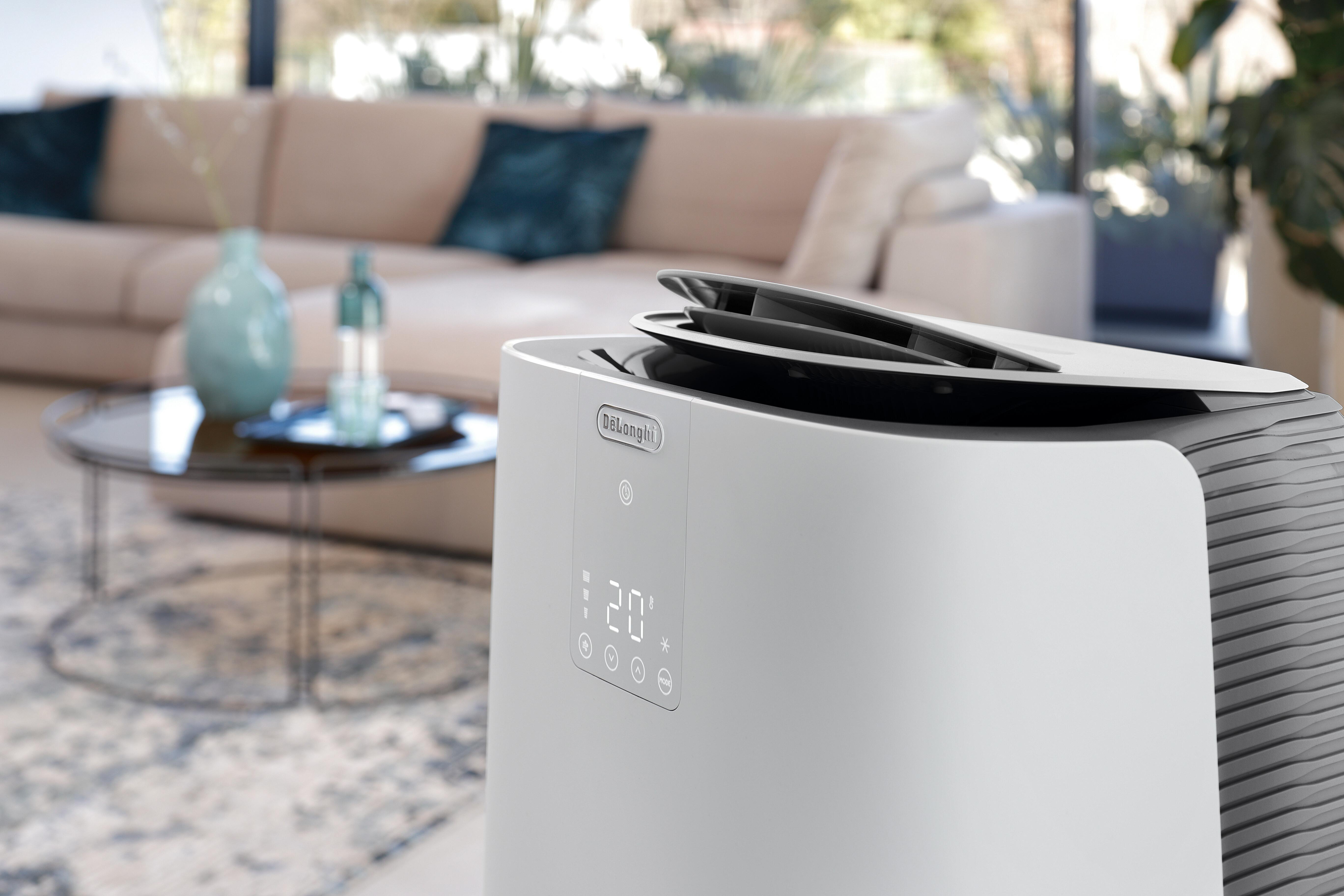 Ein weißer DeLonghi Klimaanlagen-Turm steht im Vordergrund. Im Hintergrund ein Sofa und ein Couchtisch.