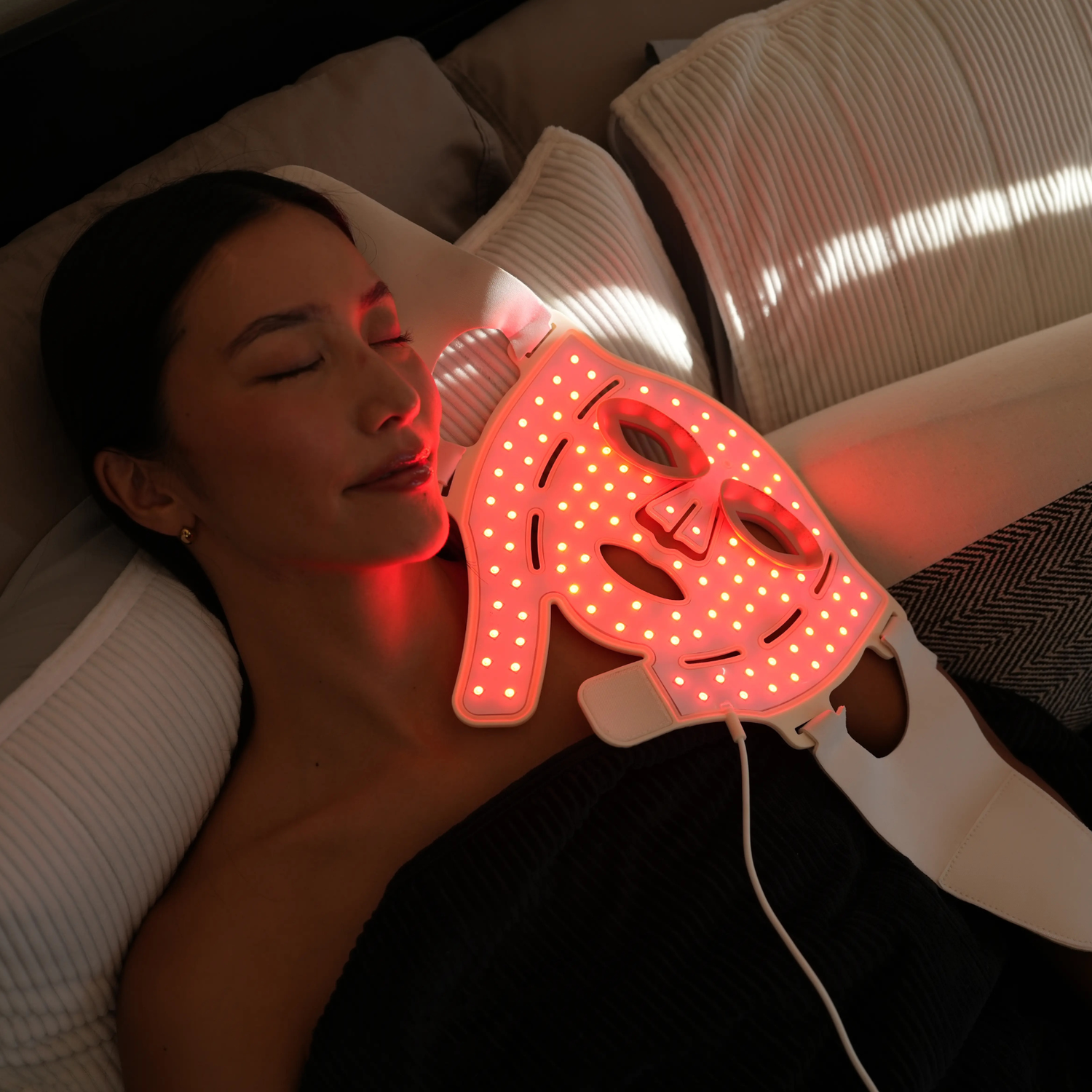 A woman lies down wearing a red LED facial mask on pillows.