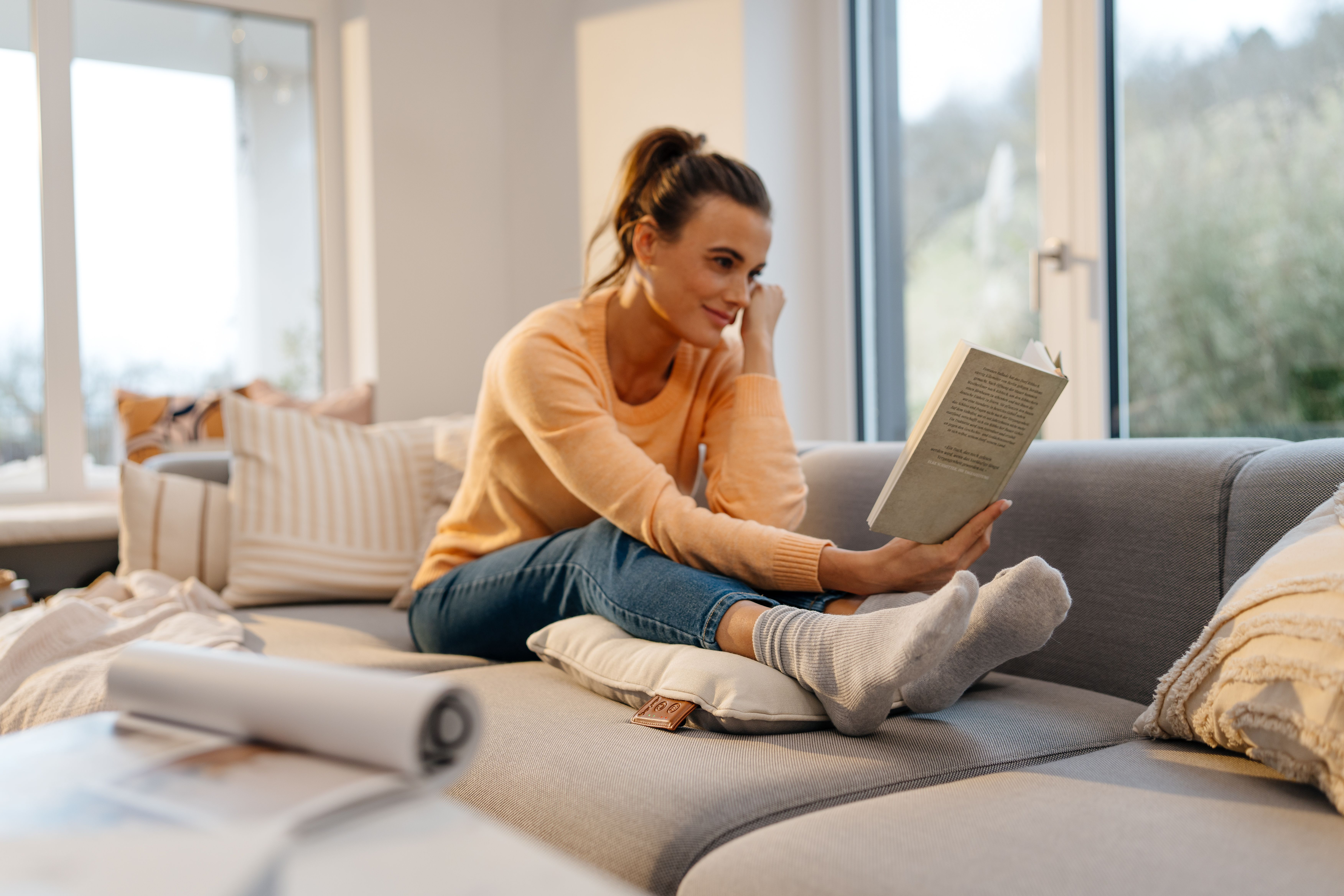 Eine Frau in einem orangefarbenen Pullover liest ein Buch auf einem grauen Sofa.