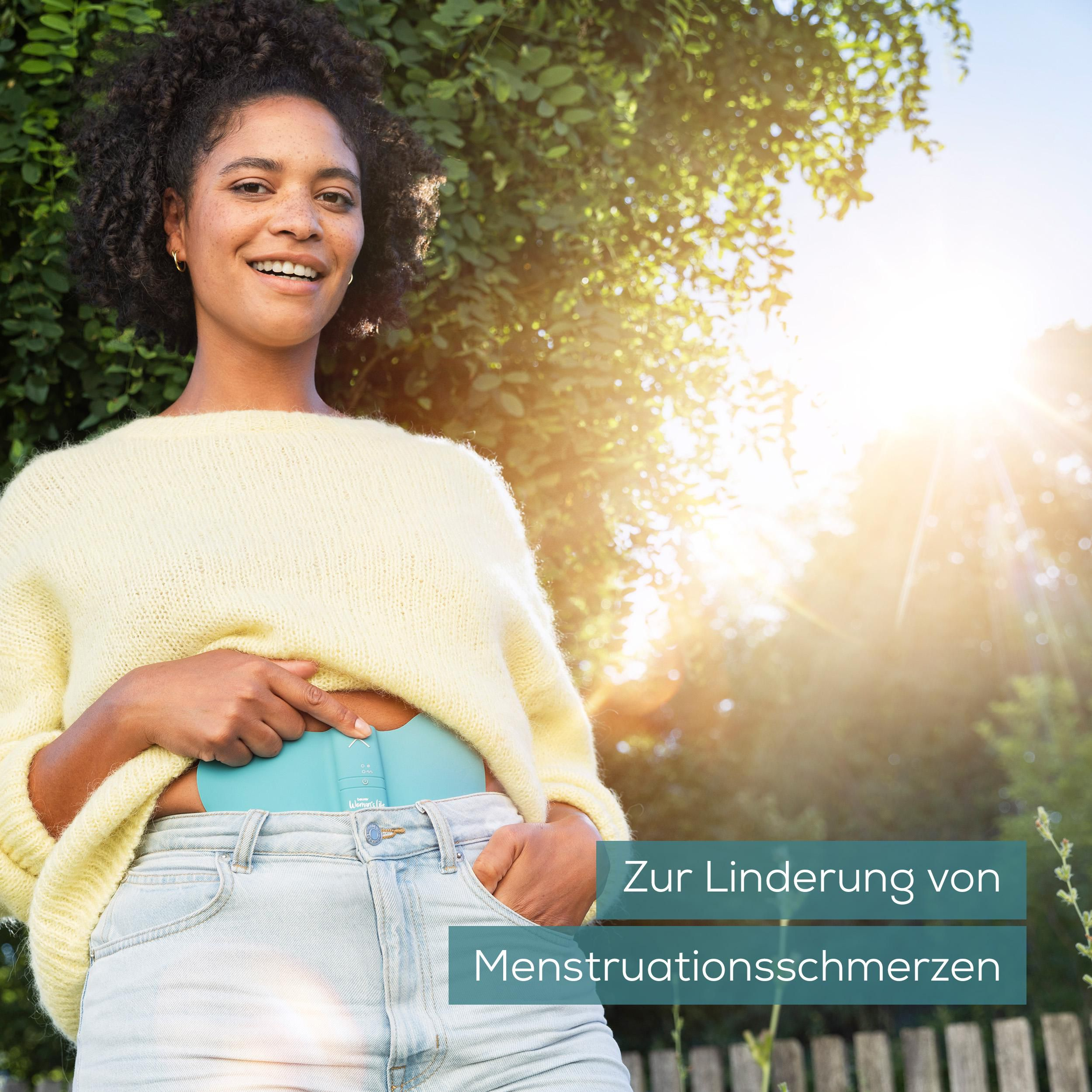 Frau mit lockigem Haar trägt hellgelben Pullover, hält blaues Gerät am Bauch.
