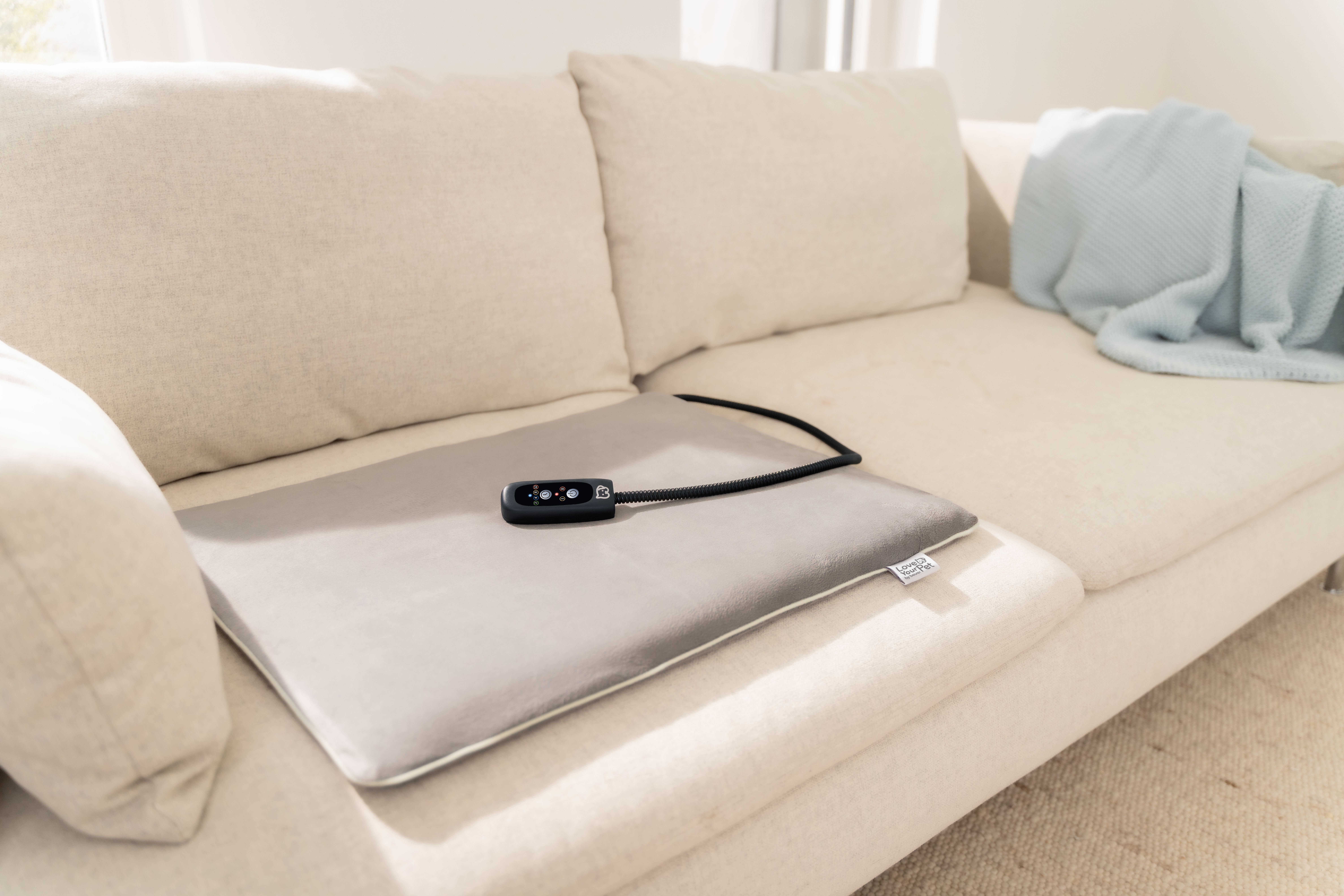Graues Heizkissen mit schwarzer Fernbedienung auf cremefarbener Couch.