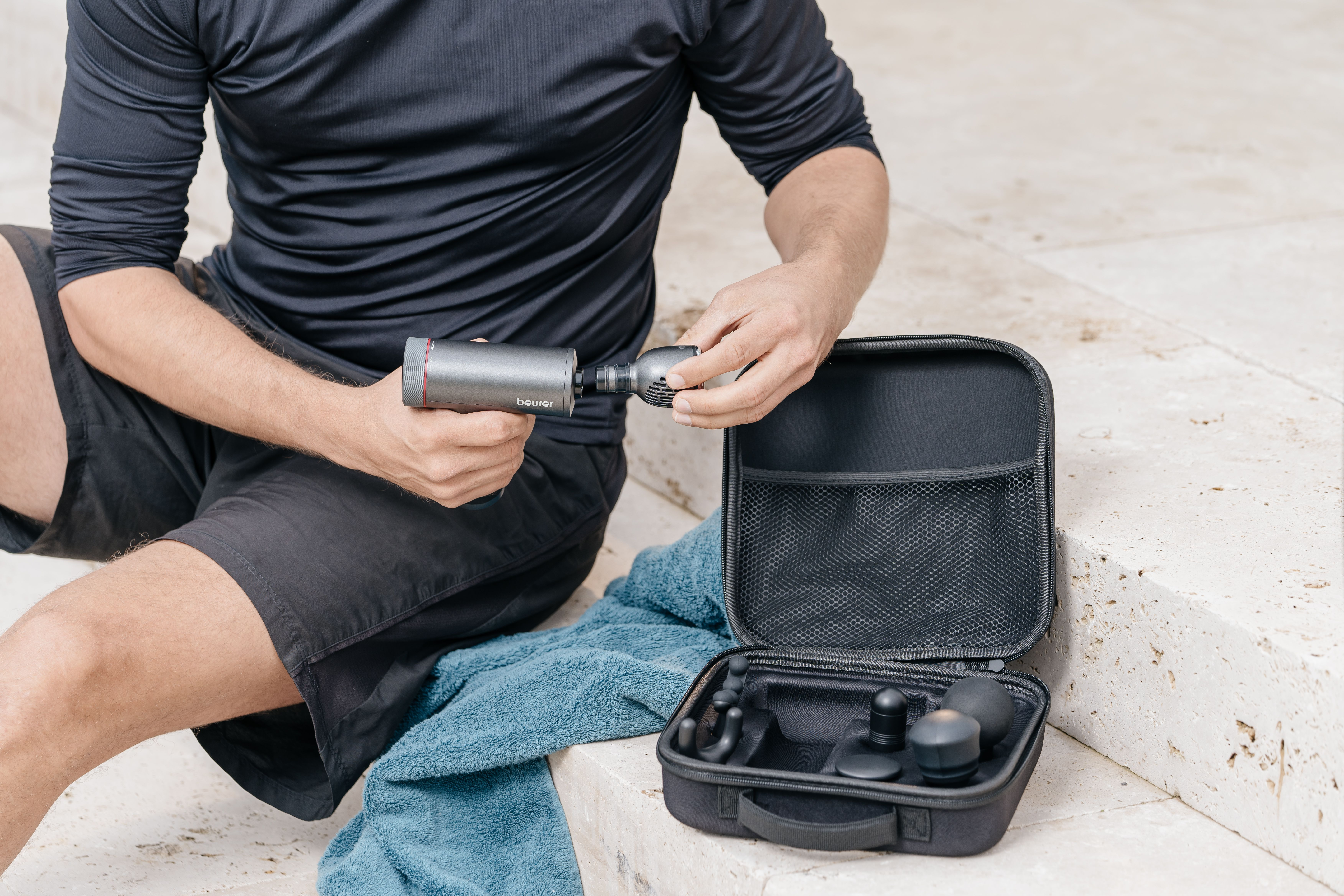 Mann in schwarzer Kleidung hantiert mit Massagepistole und Zubehör in Tasche.