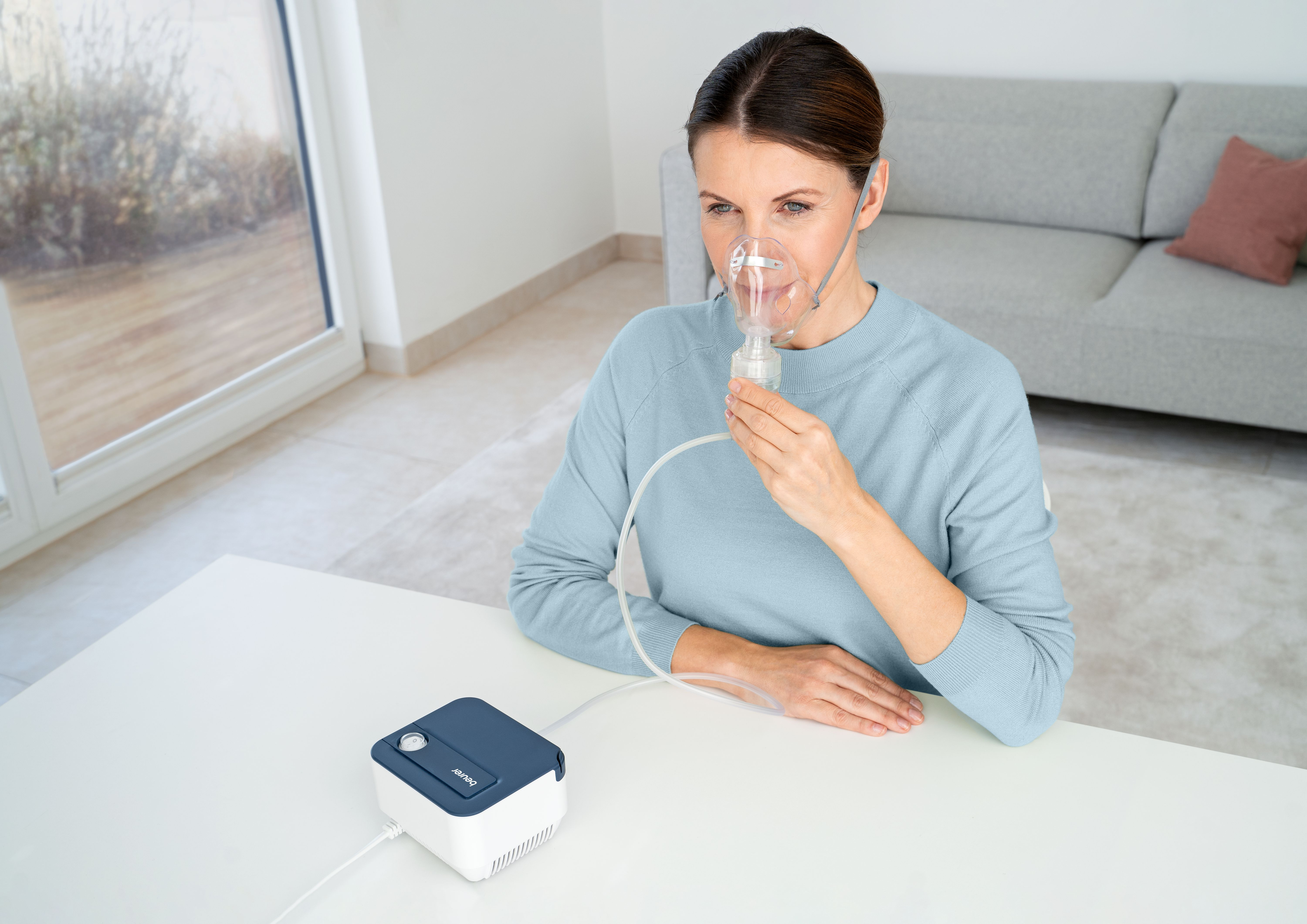 Eine Frau mit Inhalationsmaske sitzt an einem Tisch mit einem Vernebler.