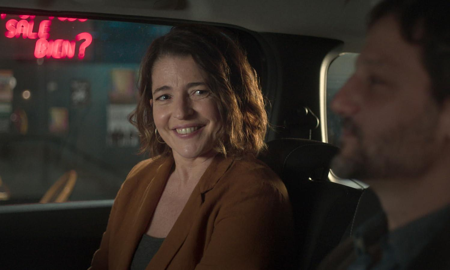 Eine Frau mit braunem Blazer lacht in einem Auto. Im Hintergrund leuchtet Neonlicht.