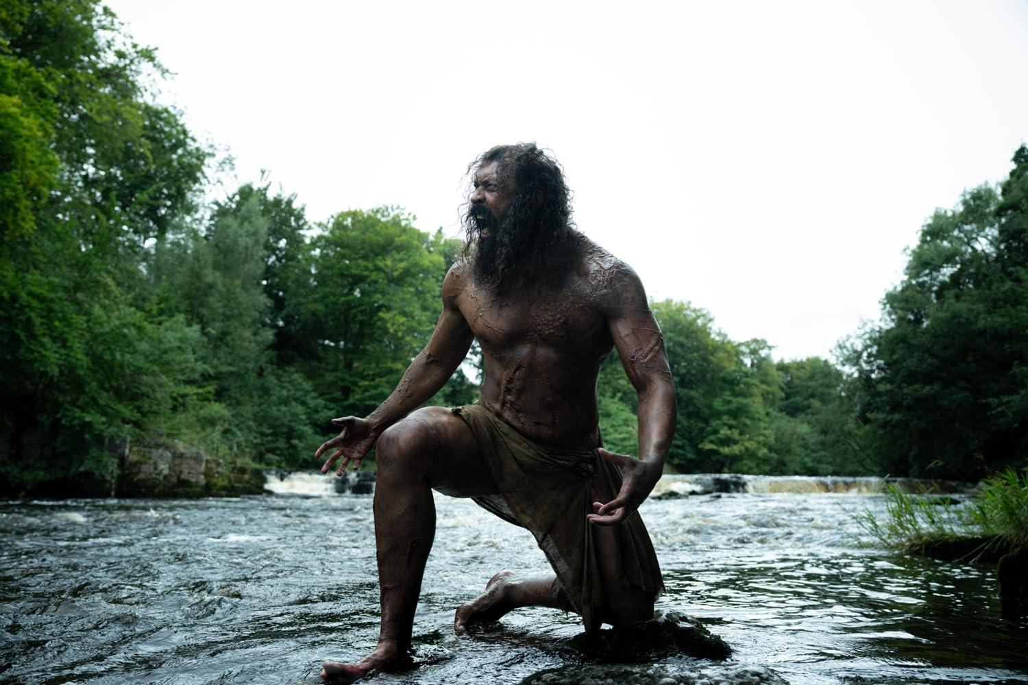 Mann mit Bart kniet in Fluss, umgeben von Bäumen.