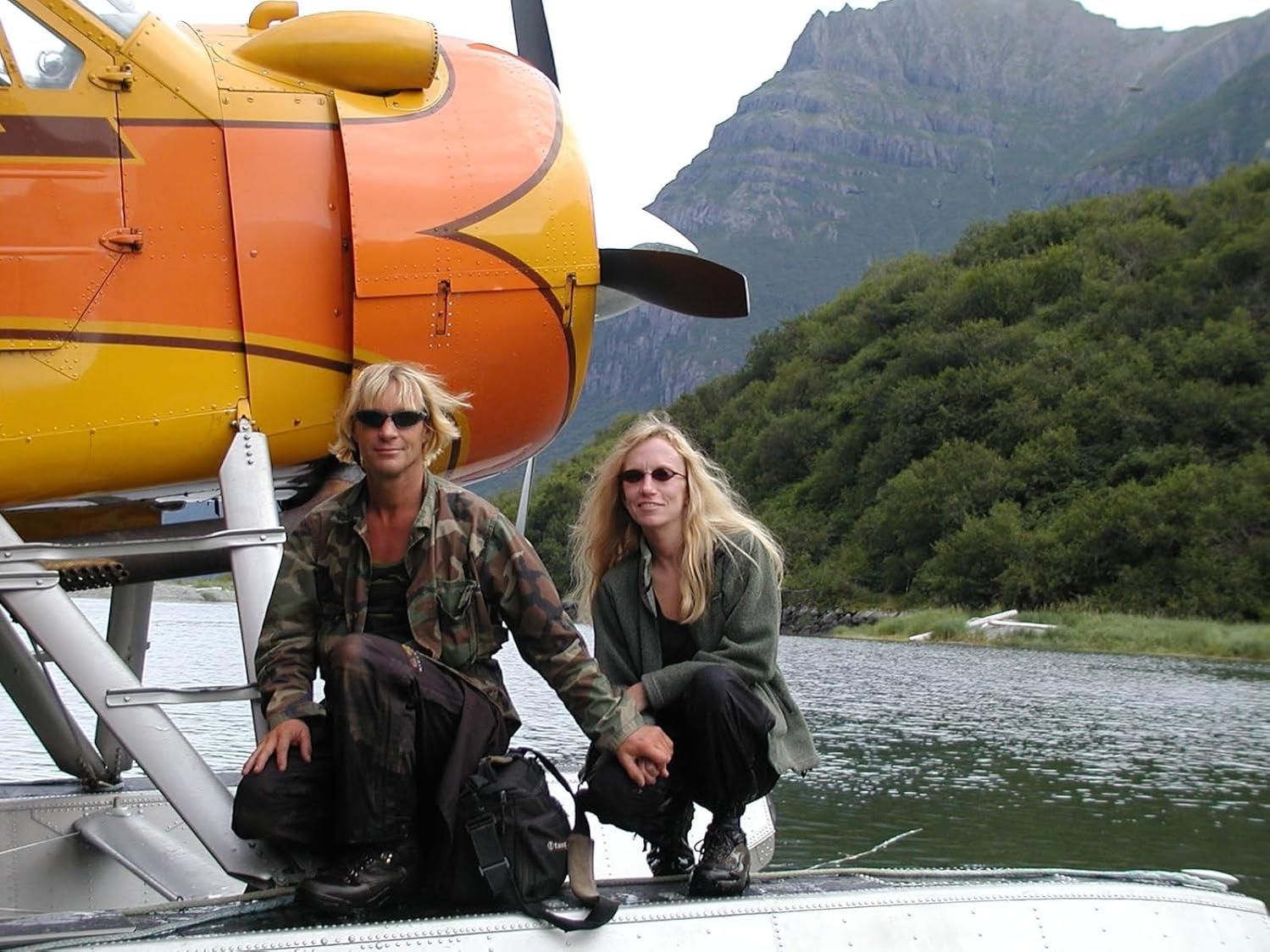Zwei Personen kauern neben einem orange-gelben Wasserflugzeug mit Bergen im Hintergrund.