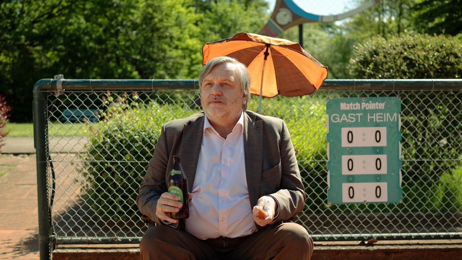 Mann mit Bierflasche und Wurst sitzt unter Sonnenschirm neben Spielstandsanzeige.