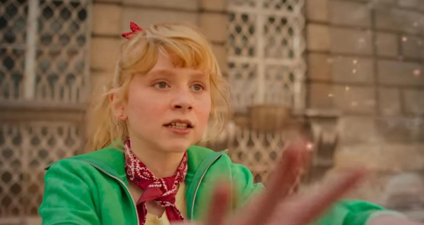 Ein junges Mädchen mit blondem Haar und einer roten Schleife im Haar, trägt eine grüne Jacke.