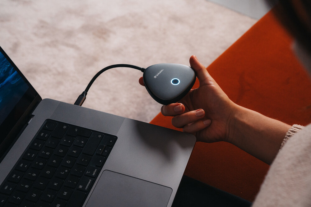 Egy kéz egy szürke USB-C adaptert tart, világító gyűrűvel egy laptop mellett.
