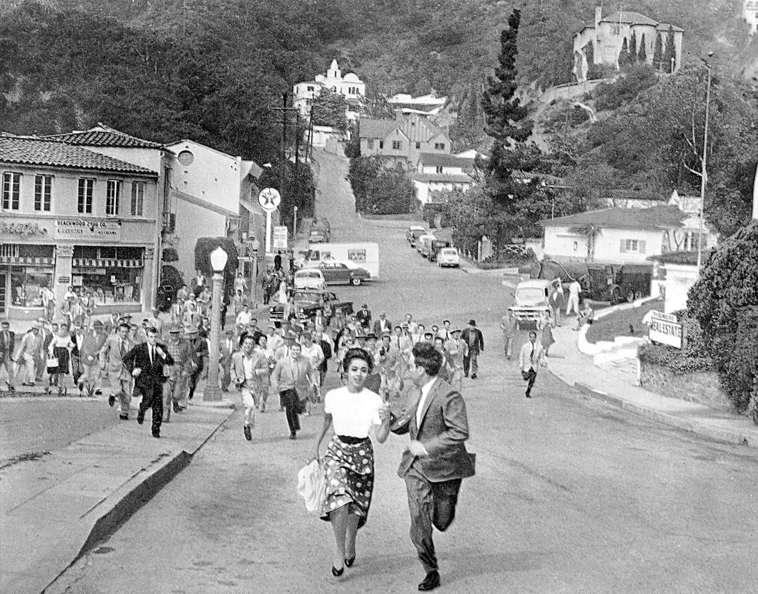 Schwarz-Weiß-Foto von Menschen, die eine Straße hinunterlaufen, mit Gebäuden und Bäumen im Hintergrund.
