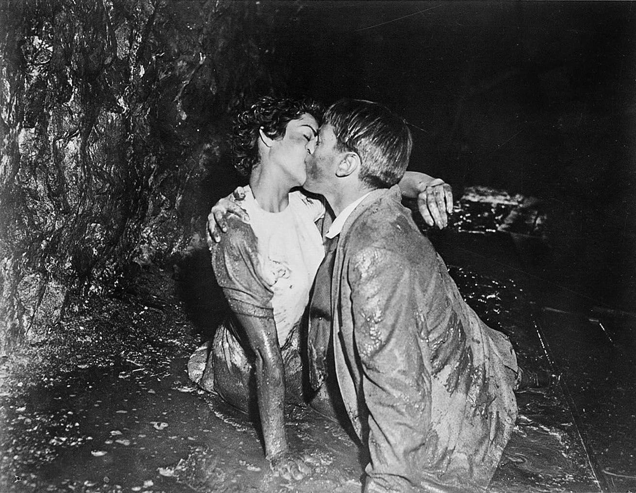Ein Schwarz-Weiß-Foto eines Paares, das sich in einer dunklen, schlammigen Höhle küsst. Beide sind mit Schlamm bedeckt.
