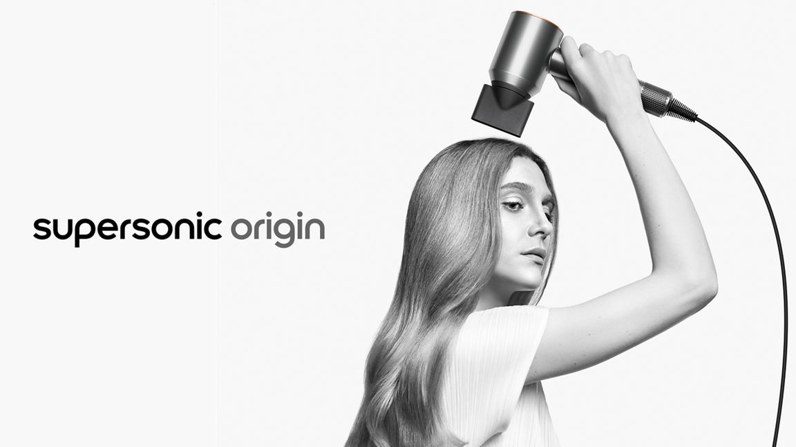 Une femme aux cheveux longs se sèche les cheveux avec un sèche-cheveux argenté sur un fond blanc. Le texte « supersonic origin » est à gauche.