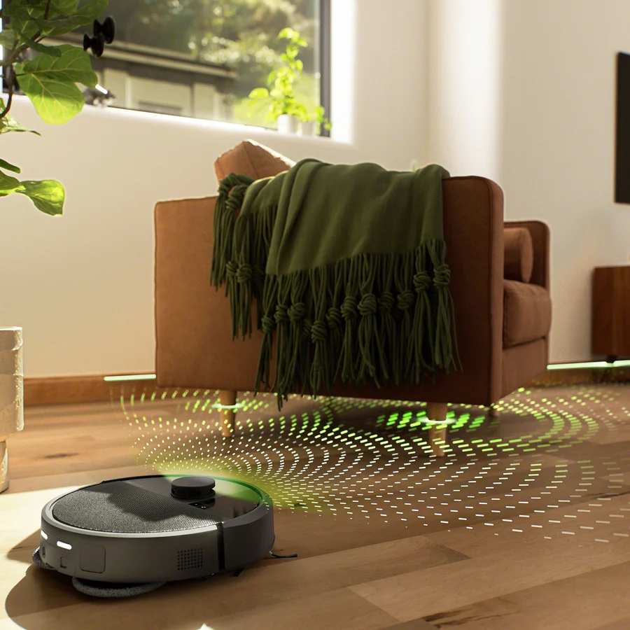 Ein Roboterstaubsauger mit grünen Lichtspuren auf einem Holzboden in einem Wohnzimmer mit braunem Sessel und grüner Decke.