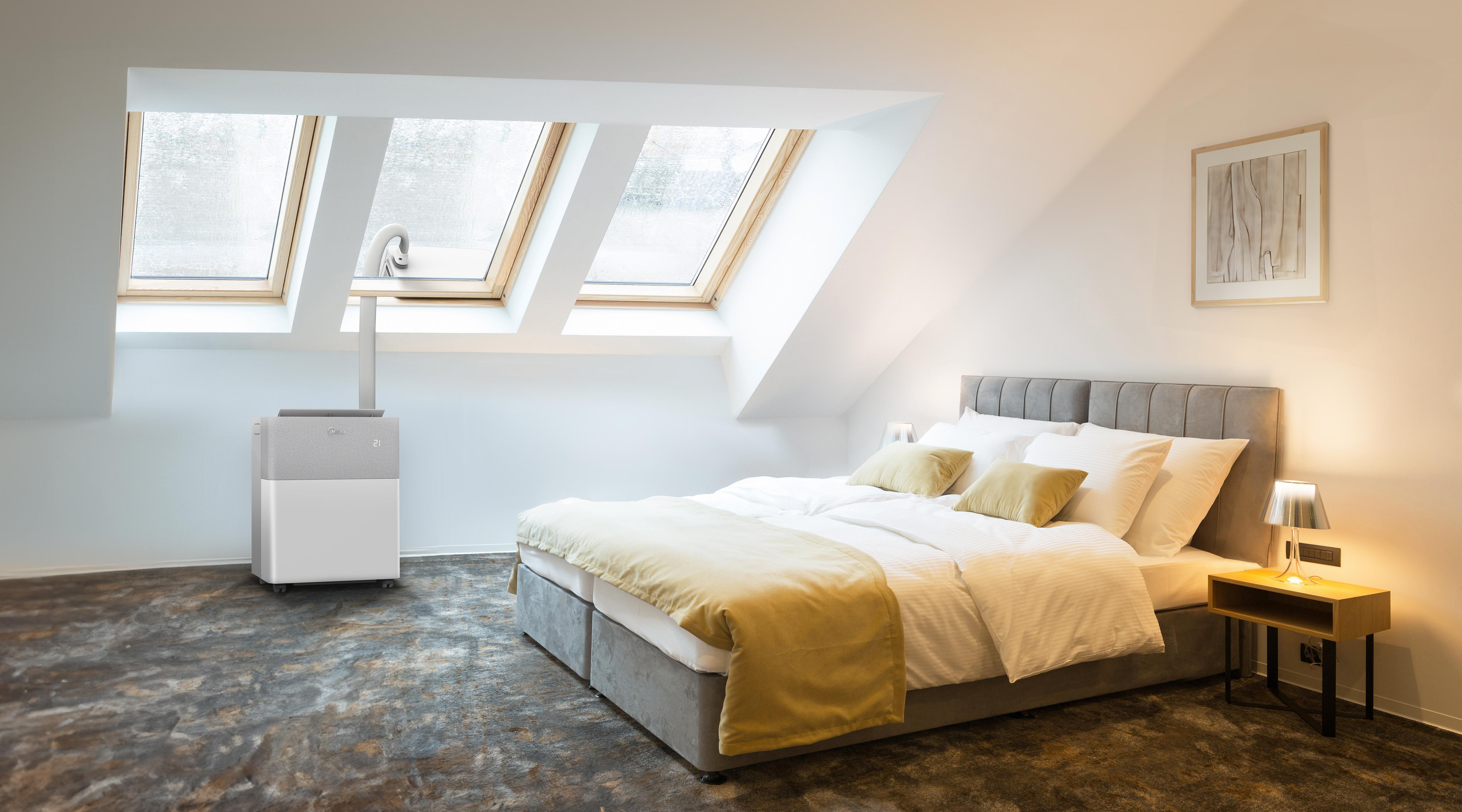 Ein weißer und grauer tragbarer Klimaanlage steht in einem modernen Schlafzimmer mit Bett, Nachttisch und drei Dachfenstern.