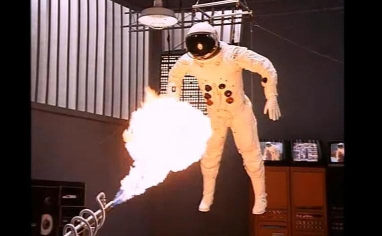 Eine Person in einem weißen Astronautenanzug schwebt, wobei ein großer Feuerball aus einem Gerät im Vordergrund hervorquillt.
