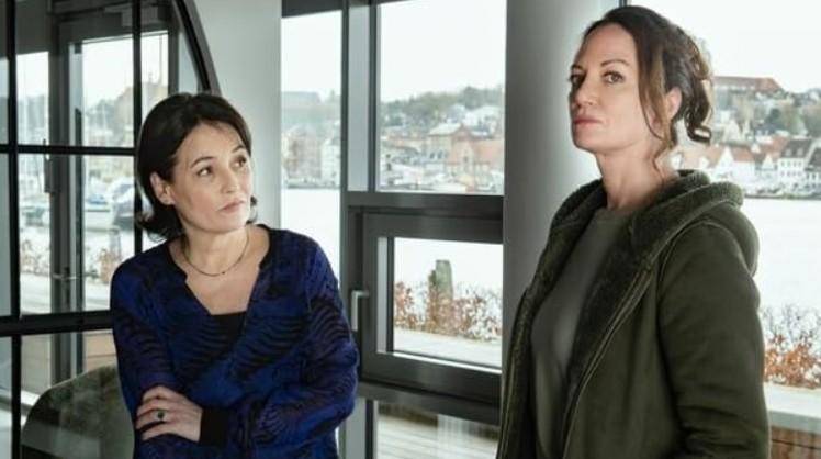 Zwei Frauen stehen in einem modernen Raum mit großen Fenstern und Blick auf eine Stadt am Wasser. Eine trägt Blau, die andere einen grünen Mantel.