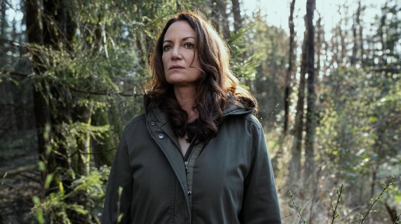 Eine Frau mit langen braunen Haaren und einer grünen Jacke steht in einem sonnigen Wald und blickt nachdenklich nach links.