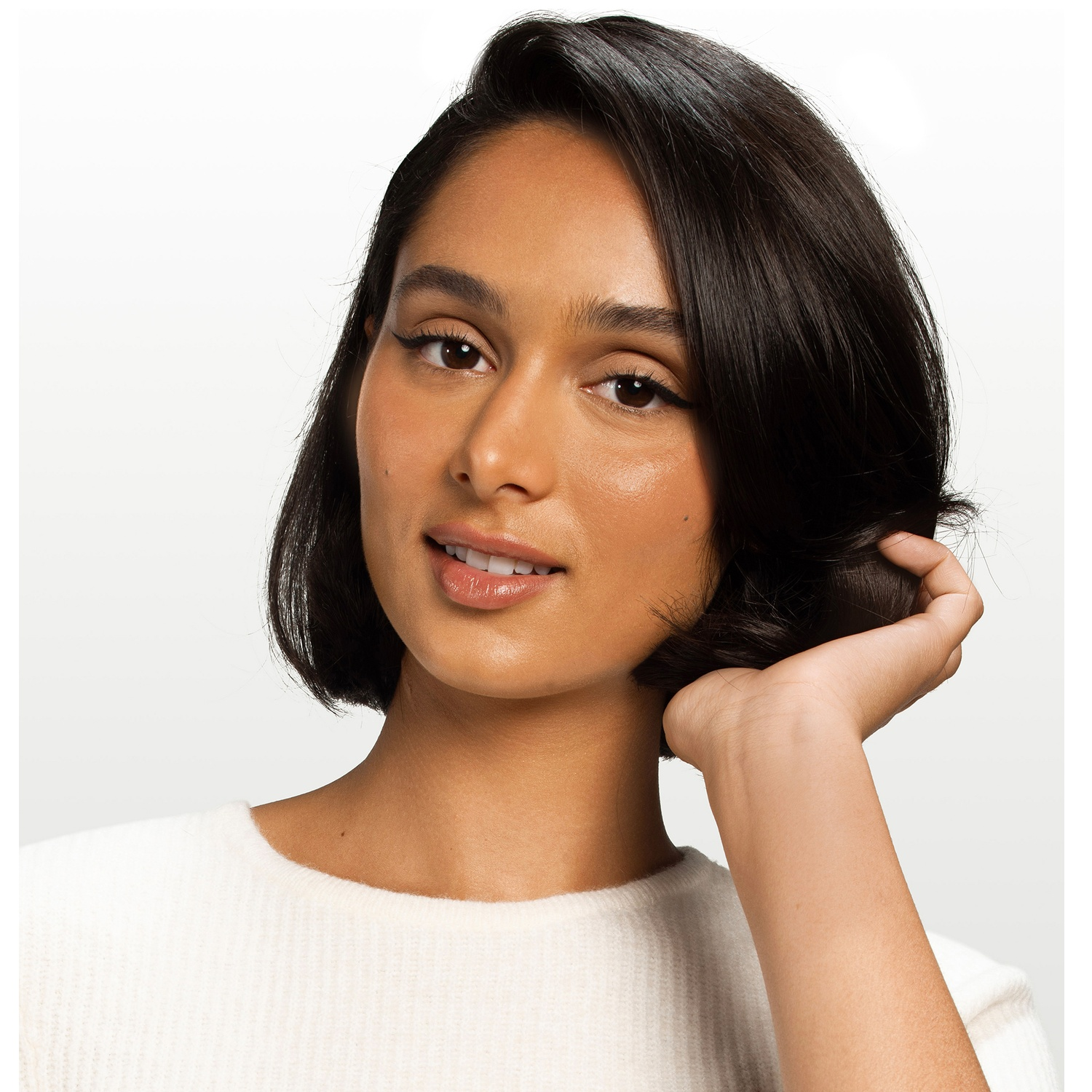 A woman with short dark hair wears a white sweater, placing her hand behind her head.