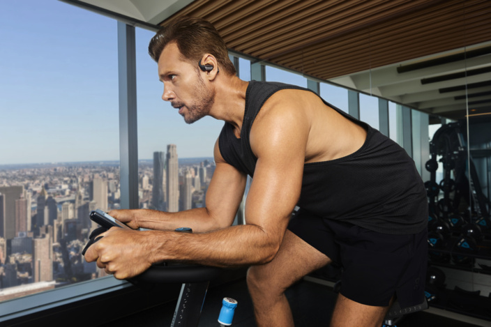 Mann auf Heimtrainer mit Kopfhörern und Handy, Stadtblick im Hintergrund.