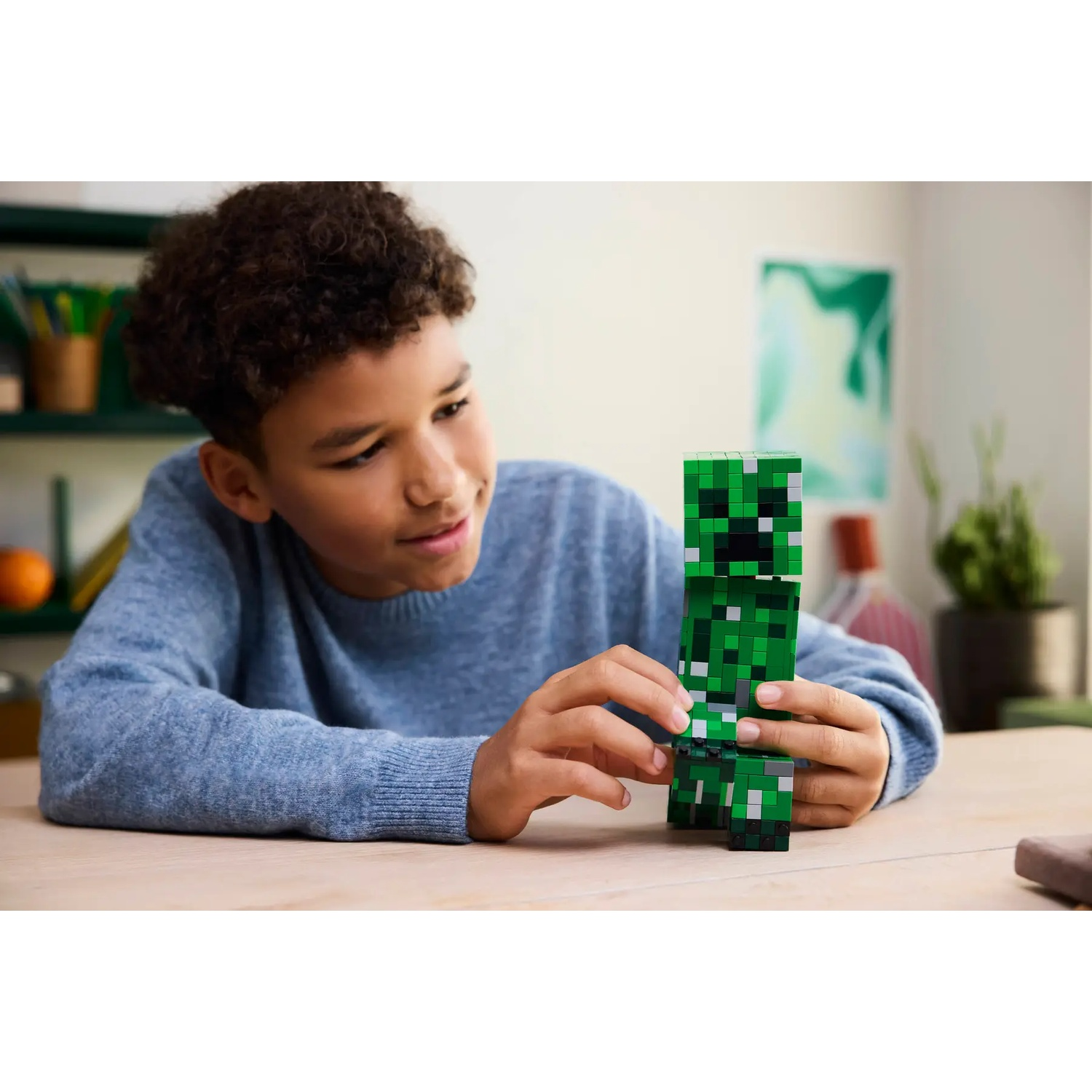 Boy builds green Minecraft figure from blocks at a wooden table.
