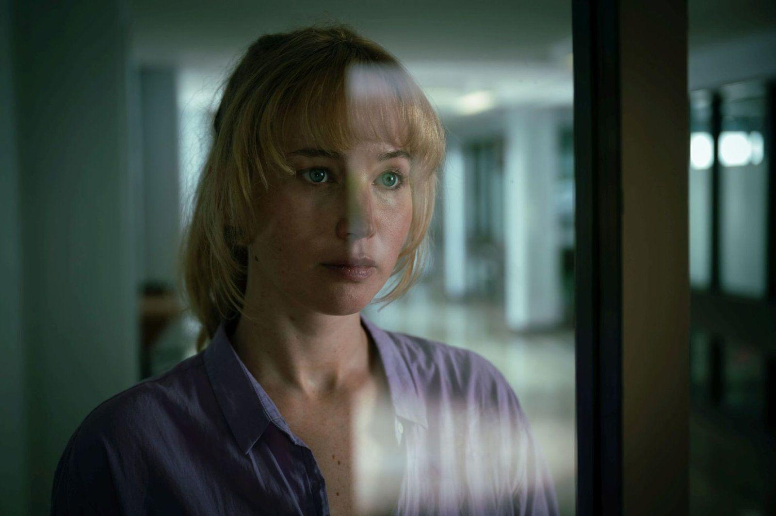 Eine Frau mit blonden Haaren und blauen Augen blickt nachdenklich durch ein Fenster.
