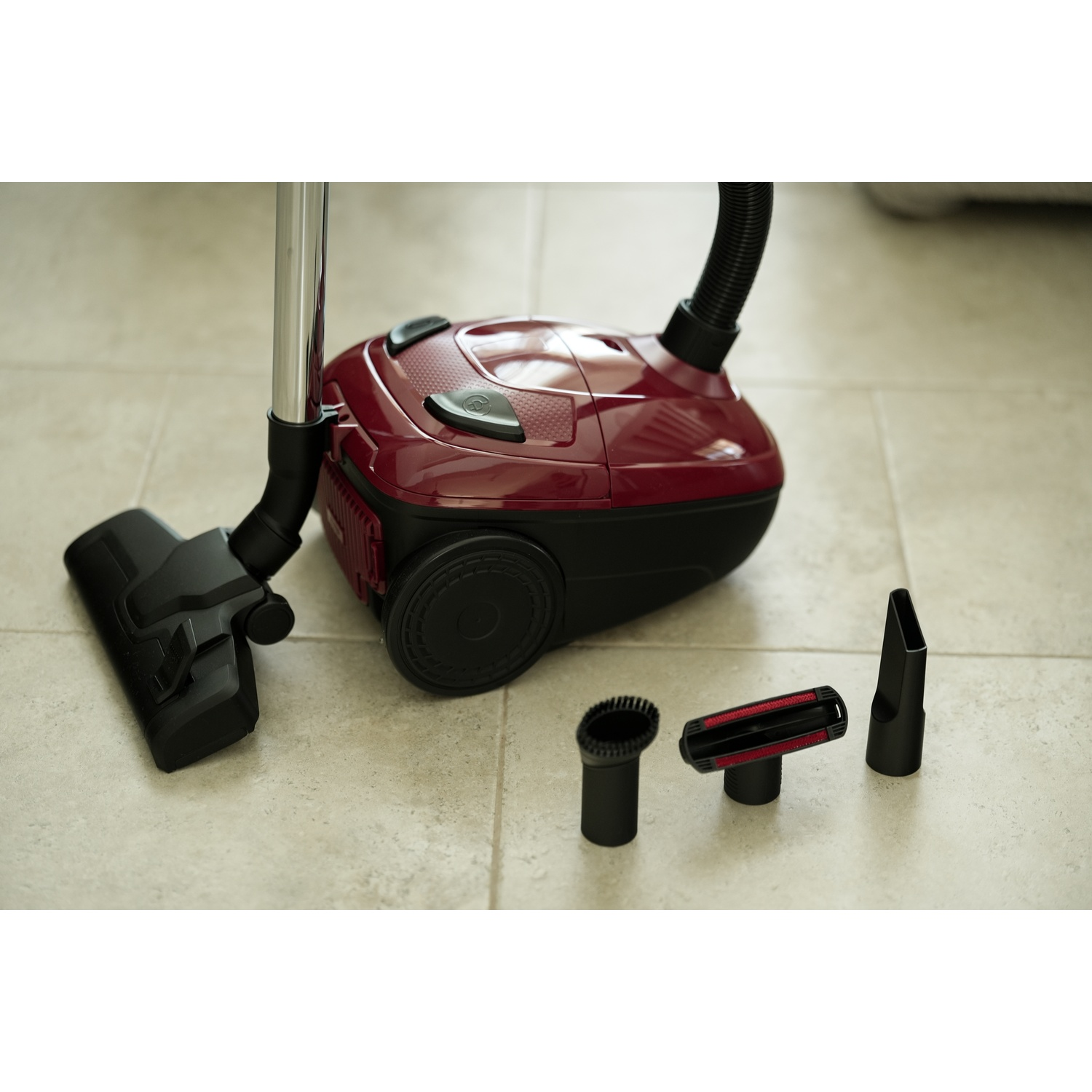 Un aspirapolvere rosso con accessori poggia su un pavimento piastrellato chiaro.