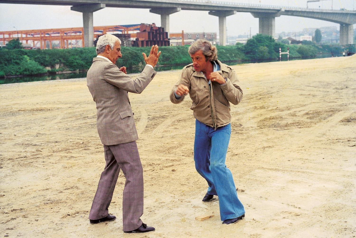 Zwei Männer in einer Kampfsportpose am Sandstrand, mit Brücke im Hintergrund.