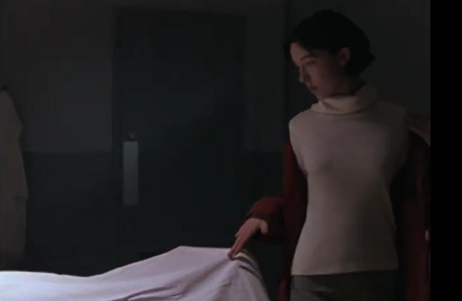Eine Frau in einem beigefarbenen Rollkragenpullover und roter Strickjacke steht neben einem Bett.