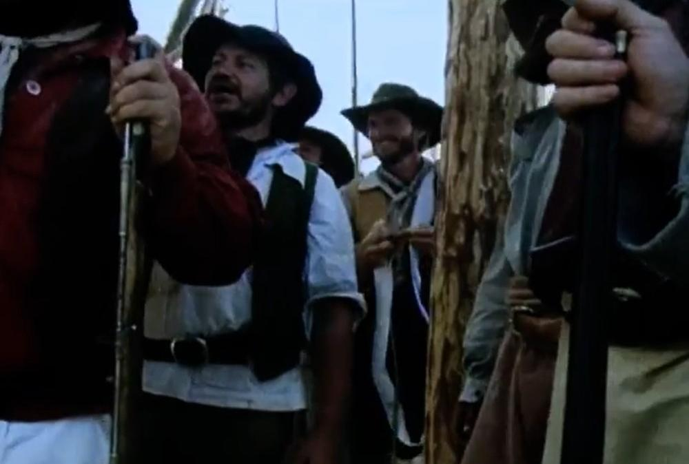 Männer in historischen Kostümen stehen eng beieinander, manche mit Hüten und Waffen.