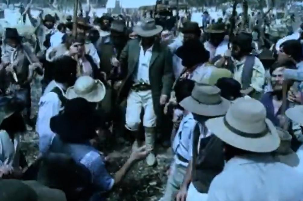Eine Gruppe von Menschen in Kleidung des 19. Jahrhunderts, viele mit Hüten, stehen eng beieinander.