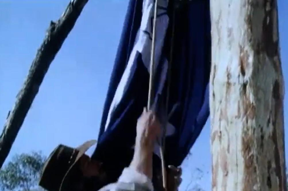 Mann in Hut hisst Flagge an Holzpfahl vor blauem Himmel.