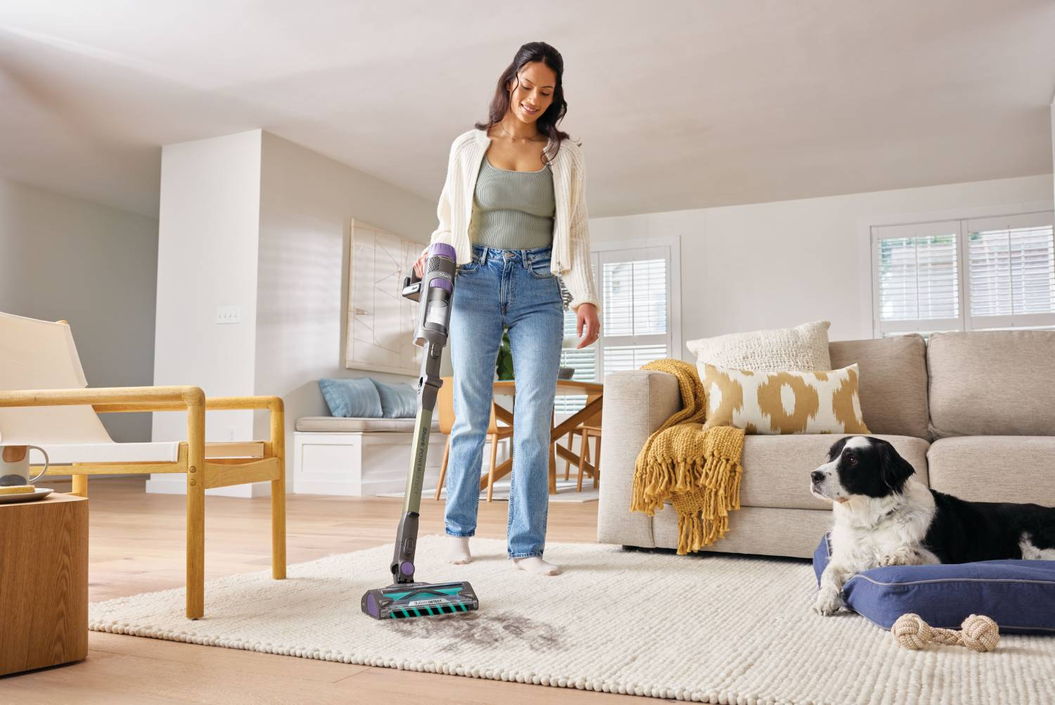 Una mujer aspira una alfombra en una sala de estar mientras un perro observa.