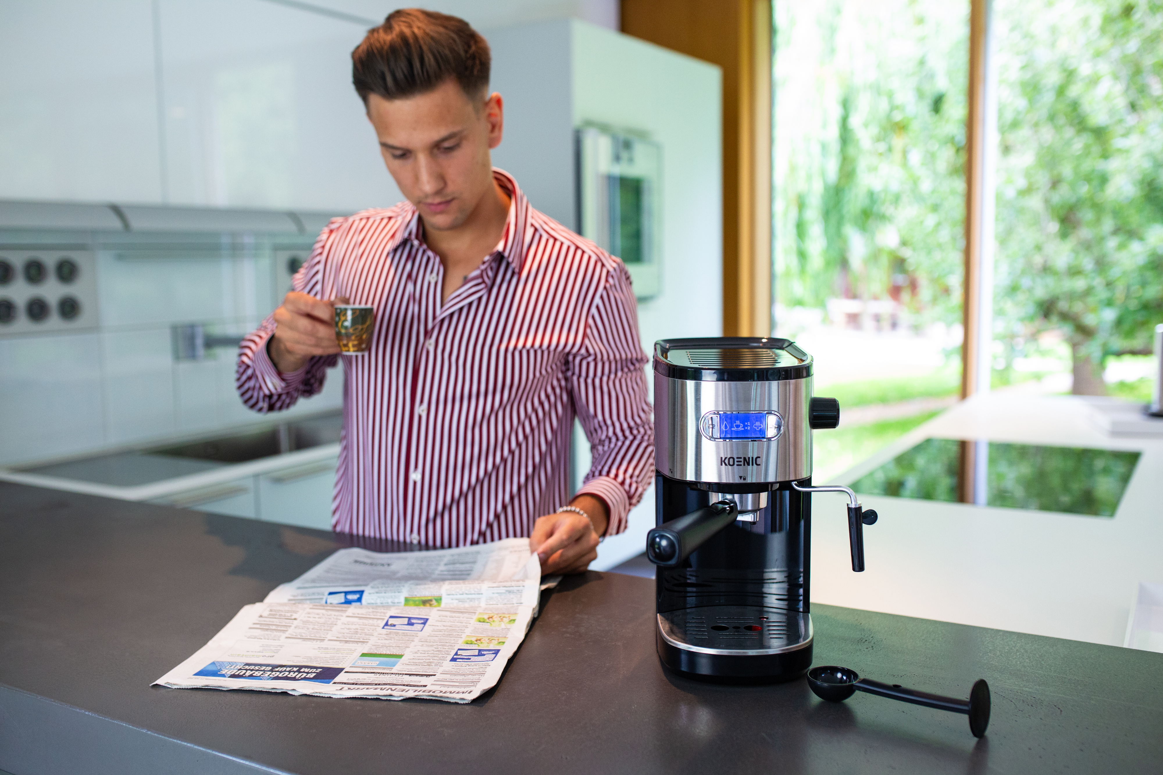 Mężczyzna w pasiastym koszuli pije kawę, czyta gazetę obok ekspresu.