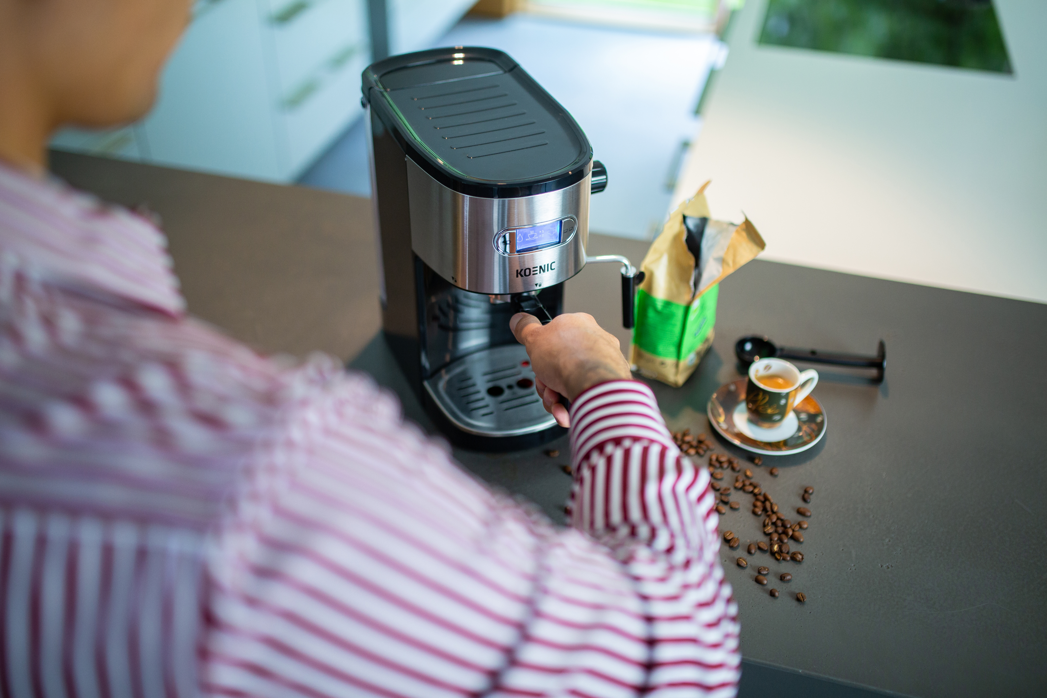Osoba w pasiastym ubraniu obsługuje ekspres do kawy. Obok stoi filiżanka espresso.