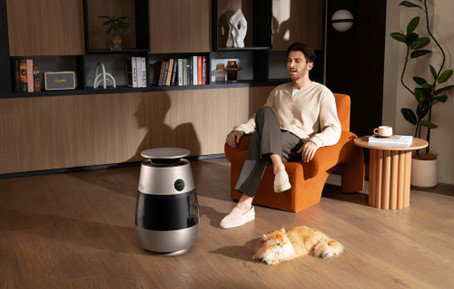 Férfi ül fotelben, macska a földön, robot és könyvespolc a háttérben.