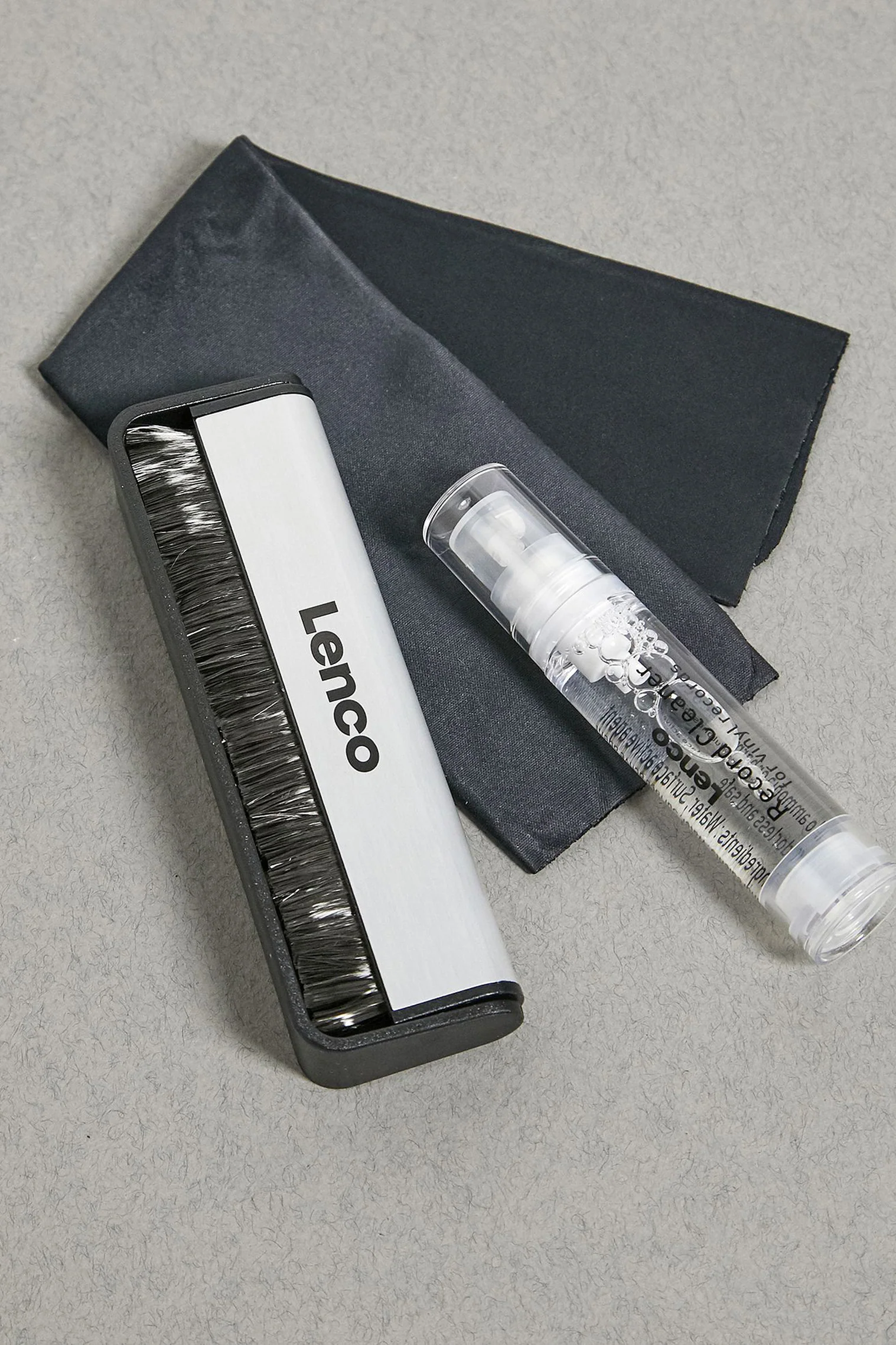 Eine Lenco Schallplattenbürste und Sprühflasche mit dunklem Tuch auf grauer Oberfläche.
