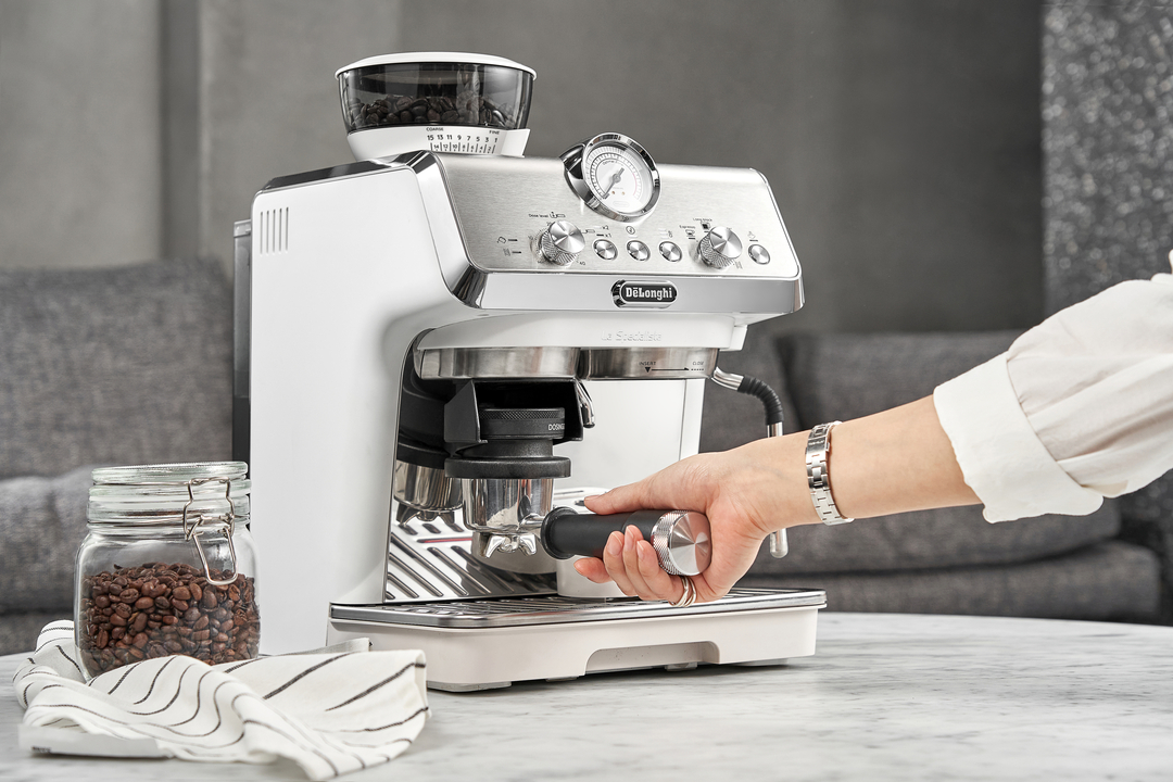 Eine Hand bedient eine weiße Espressomaschine mit Kaffeebohnenglas daneben.