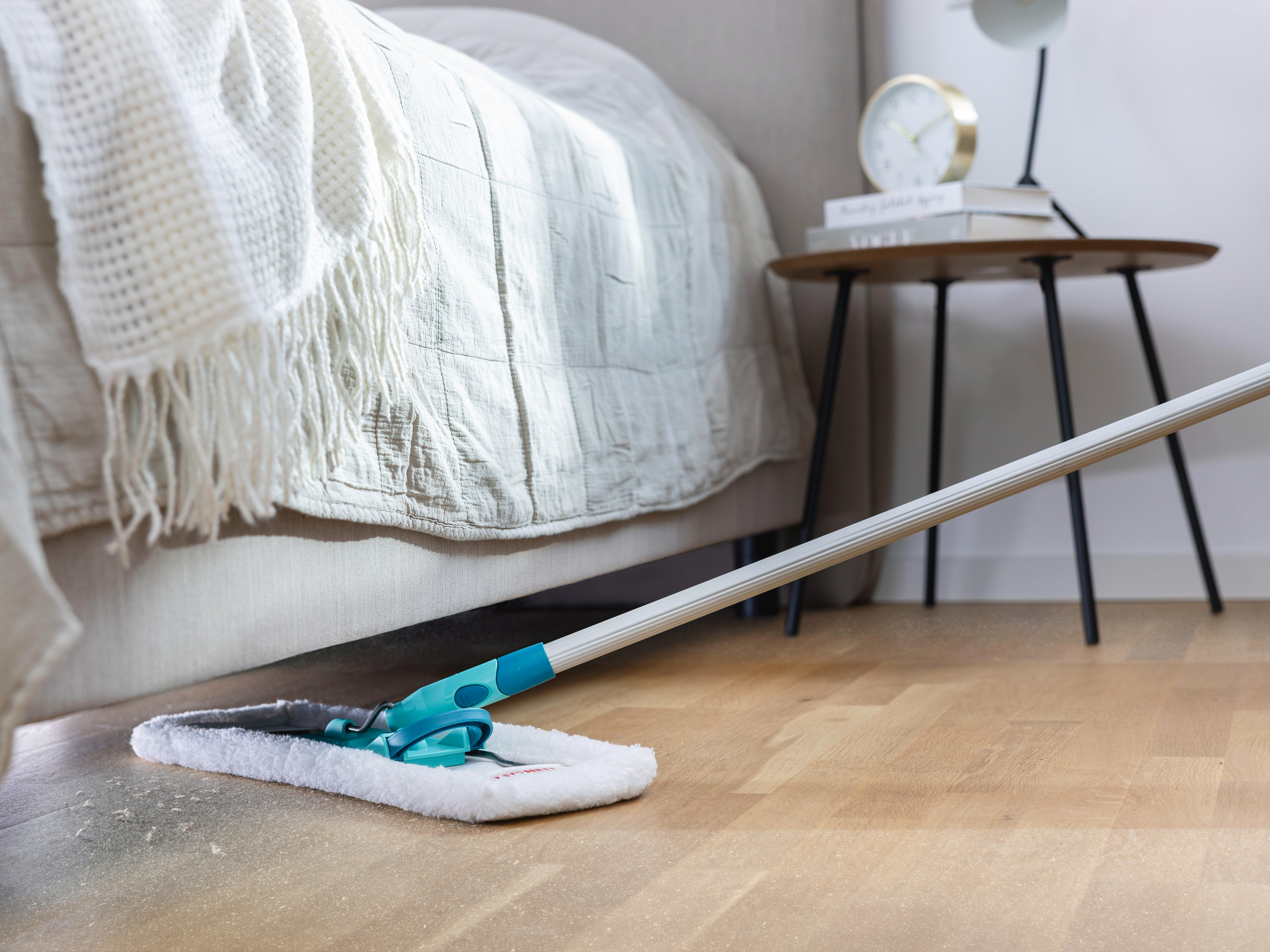 Ein Mopp reinigt einen staubigen Holzboden neben einem Bett mit weißer Decke.
