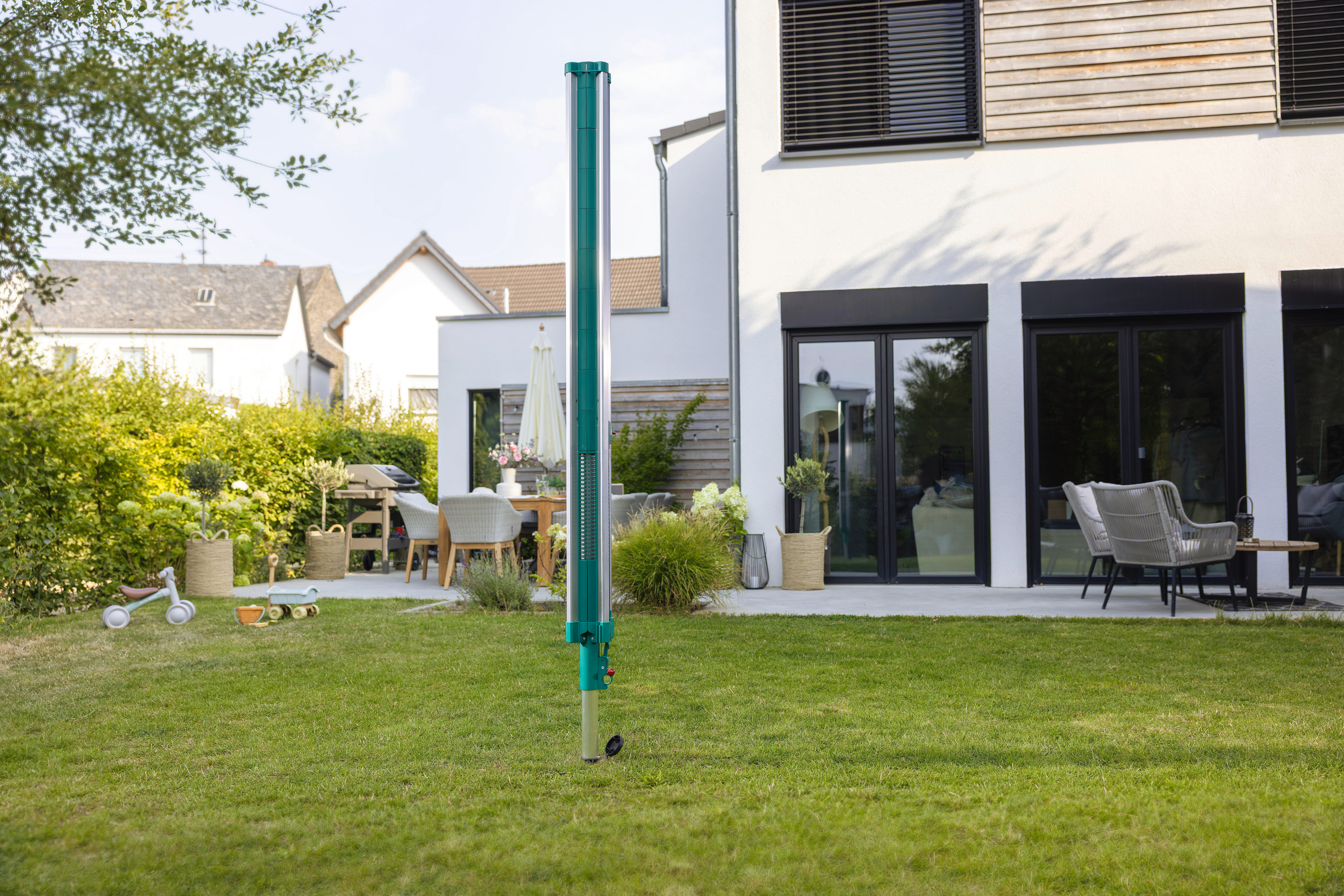 Ein grüner Messmast steht auf einem Rasen vor einem Haus mit Gartenmöbeln.