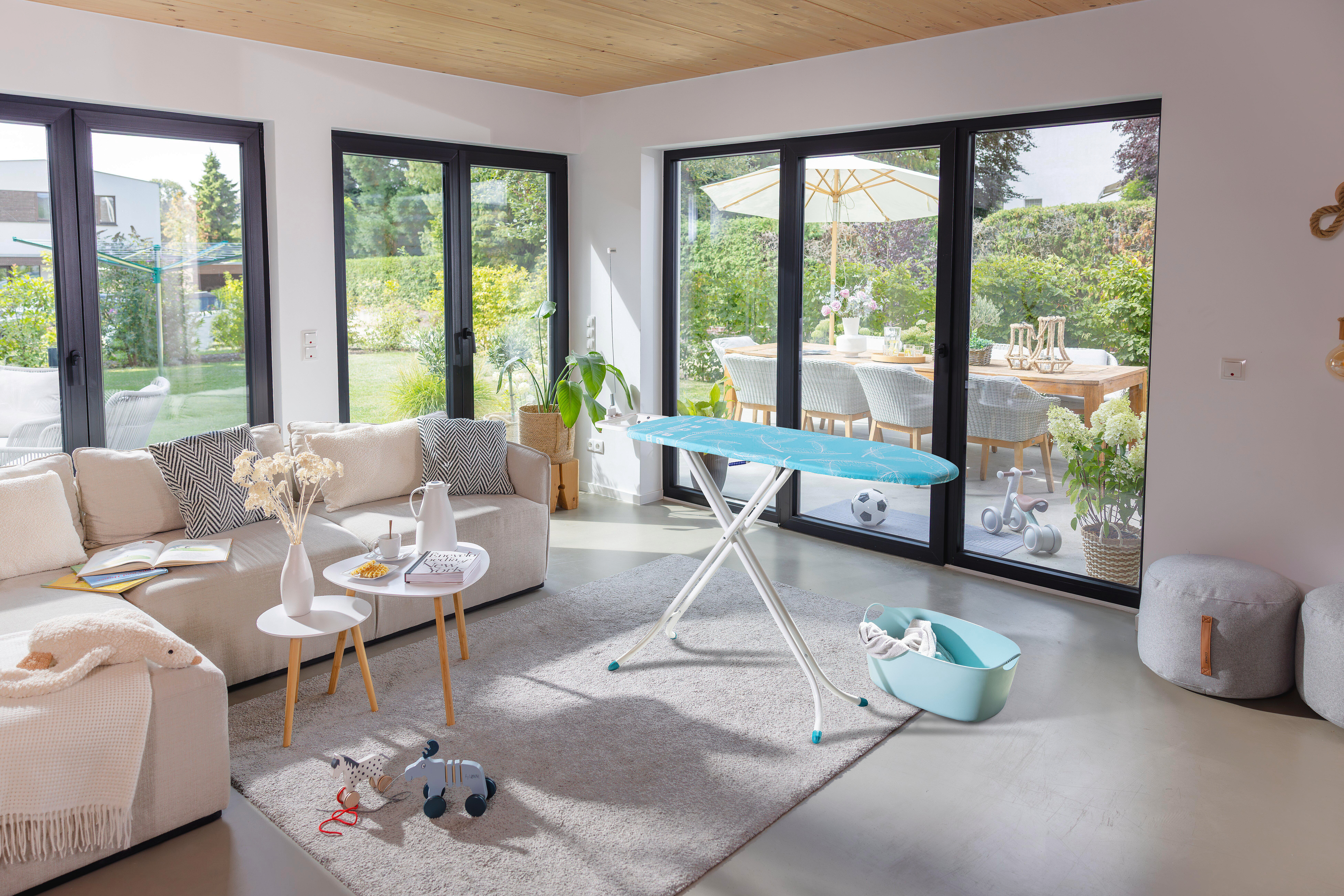 Ein Wohnzimmer mit Sofa, Bügelbrett und Blick auf einen Garten mit Esstisch.