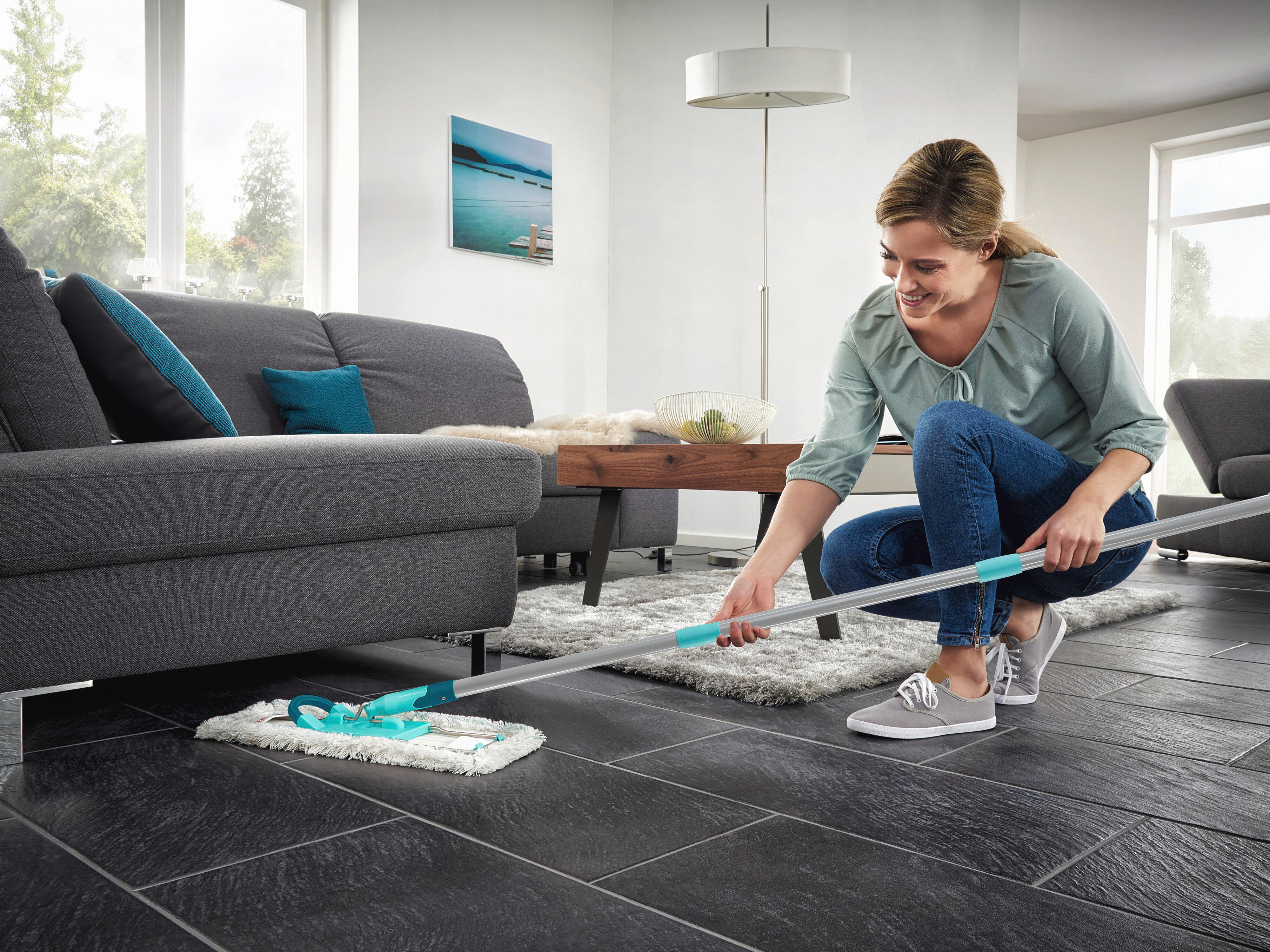 Eine Frau wischt den Boden in einem modernen Wohnzimmer mit grauer Couch.
