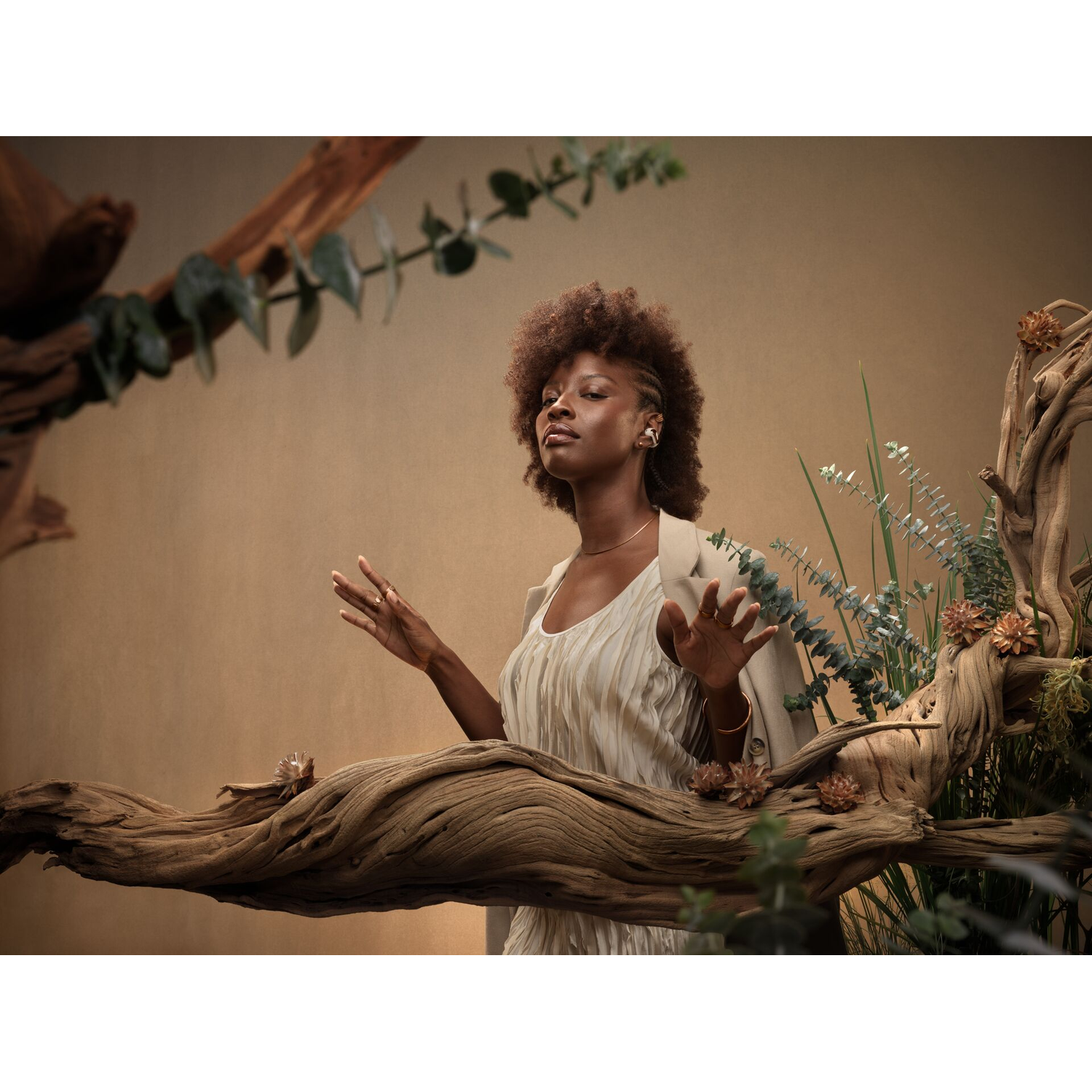 Woman with dreadlocks in dress and jacket stands between wood and plants with eyes closed.