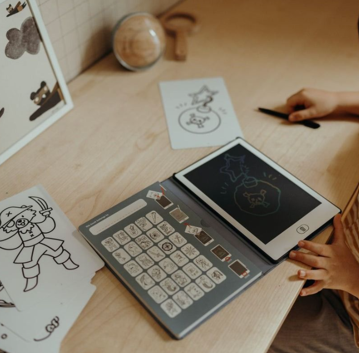 Kinderhände malen einen Piraten-Schädel auf ein Tablet. Piratenzeichnungen und ein Nachschlagewerk liegen daneben.