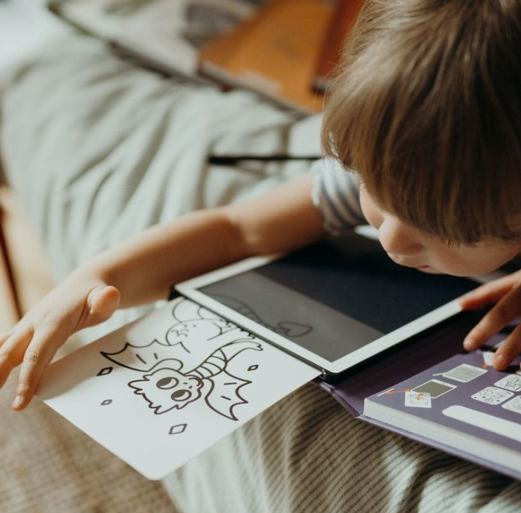 Ein Kind liegt auf einem Bett und betrachtet ein Tablet und ein Malbuch mit einem Drachen.