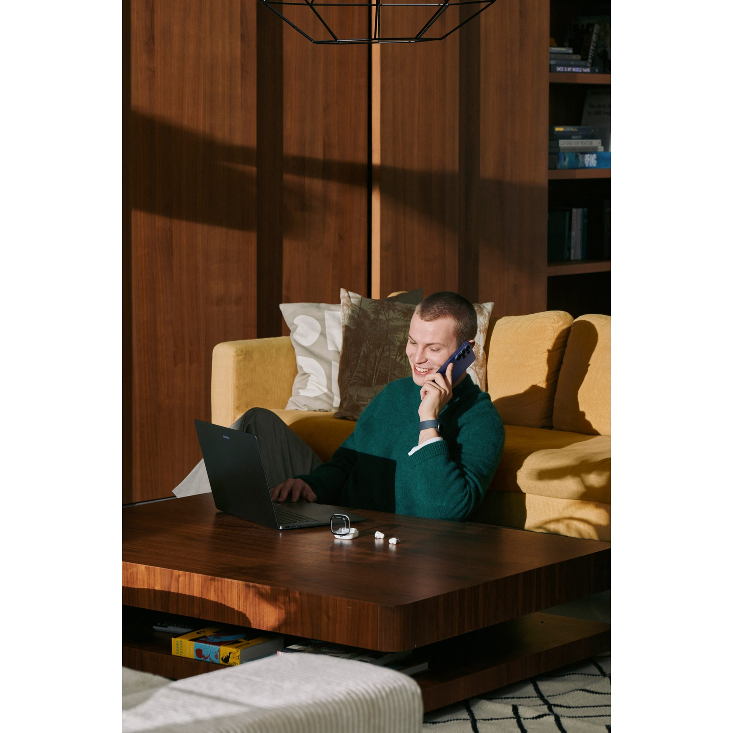 Man on yellow couch talking on phone with laptop and earbuds on coffee table.