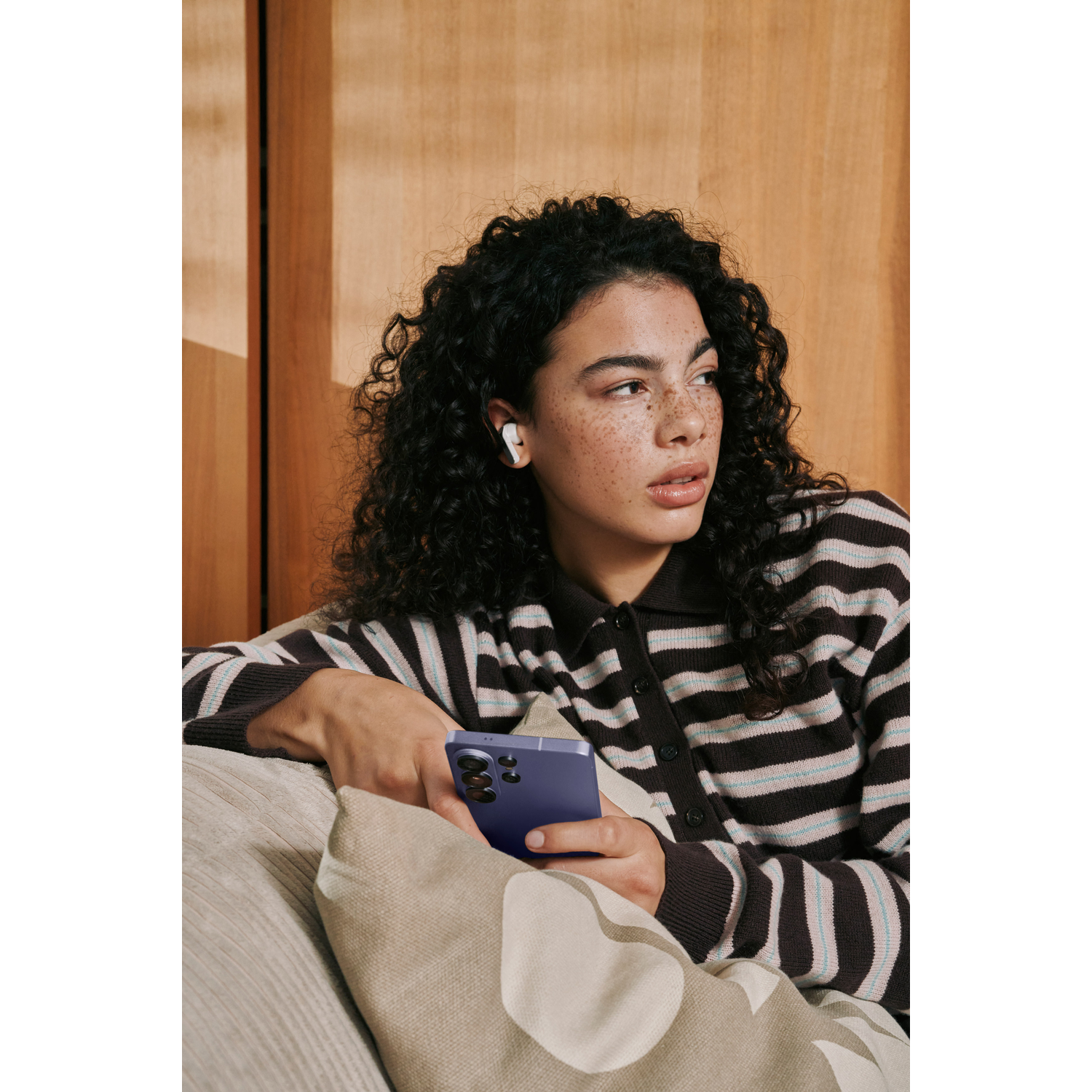 Vrouw met krullend haar draagt gestreepte vest en houdt een paarse telefoon vast.