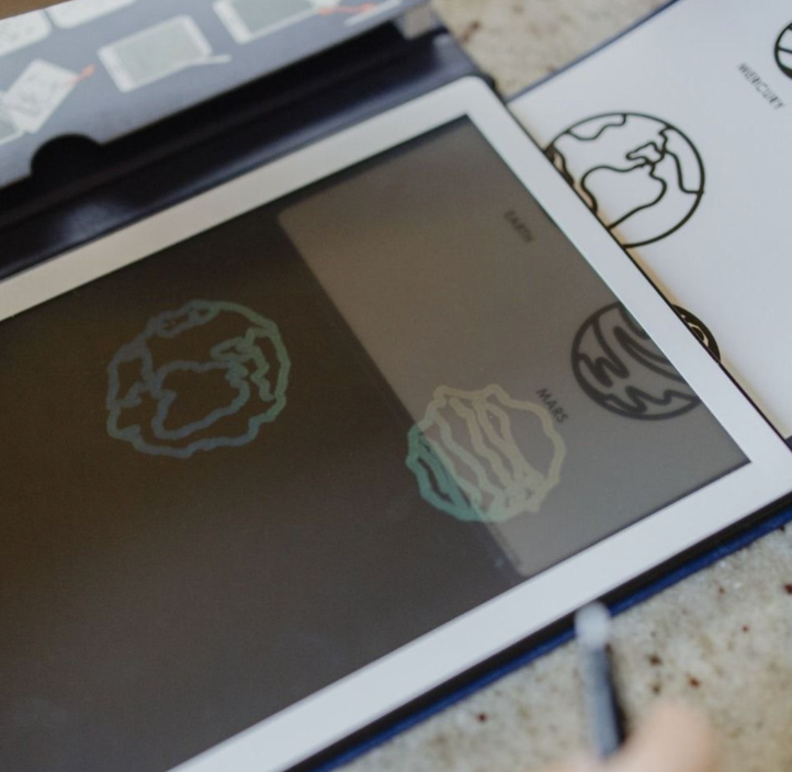 Ein Tablet zeigt eine gezeichnete Erde und den Mars. Daneben liegt ein Blatt mit Planetenbildern.