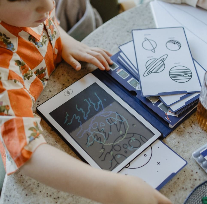 Ein Kind mit einem orange-karierten Hemd lernt mit einem Tablet und Sternkarten.