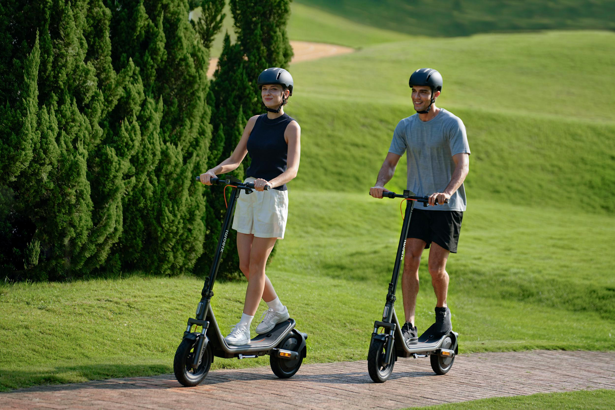 Zwei Personen mit Helmen fahren E-Scooter auf einem Weg im Grünen.